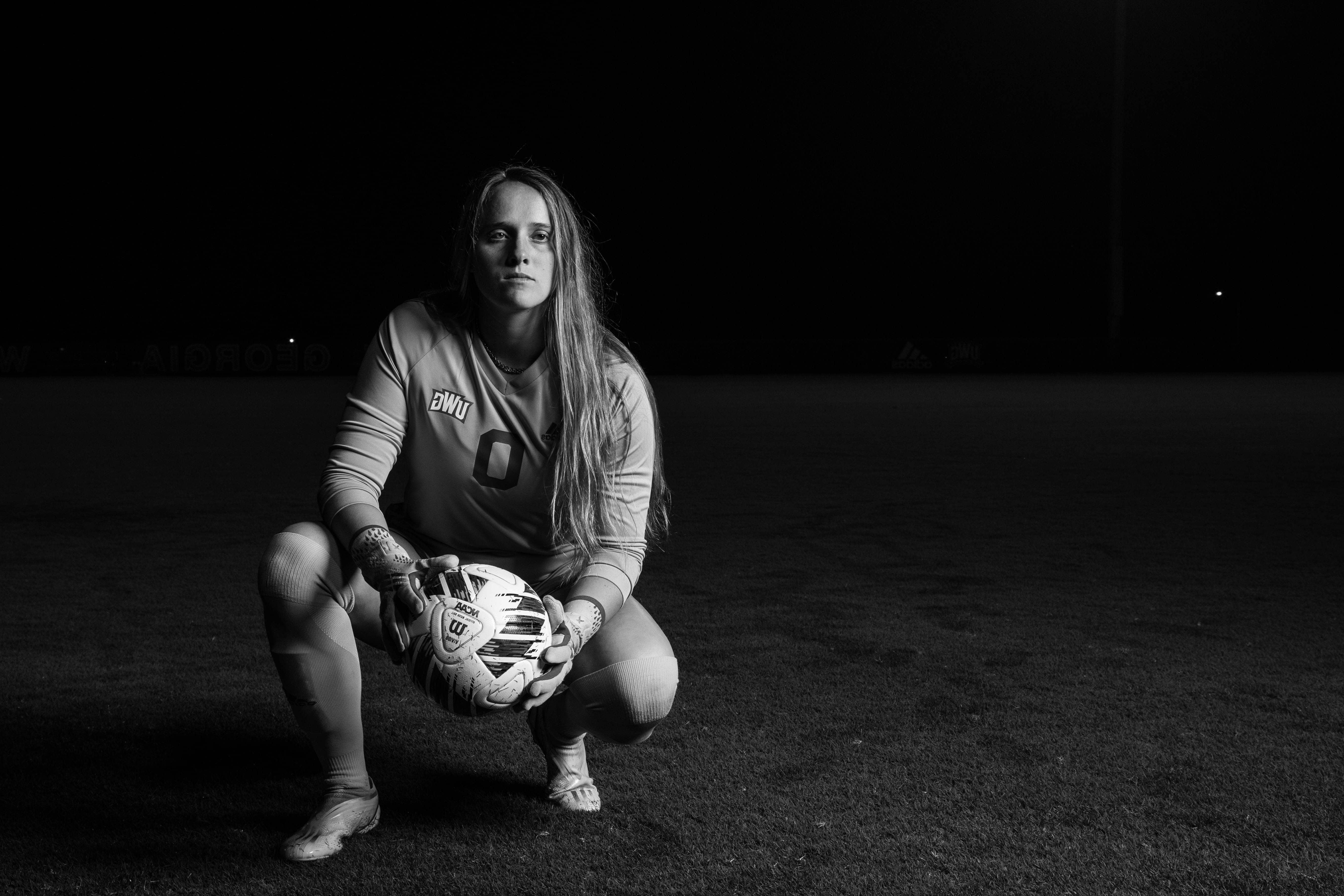 博彩平台推荐 Women's Soccer player kneeling on the field holding a soccer ball. 